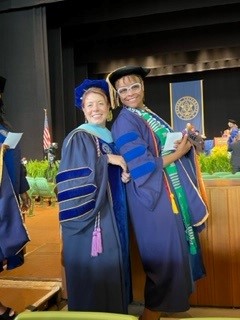Two women wearing doctoral graduation robes and caps standing in front of a stage.